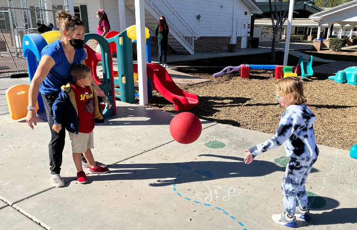 Woman helping child bounce a ball to another child.